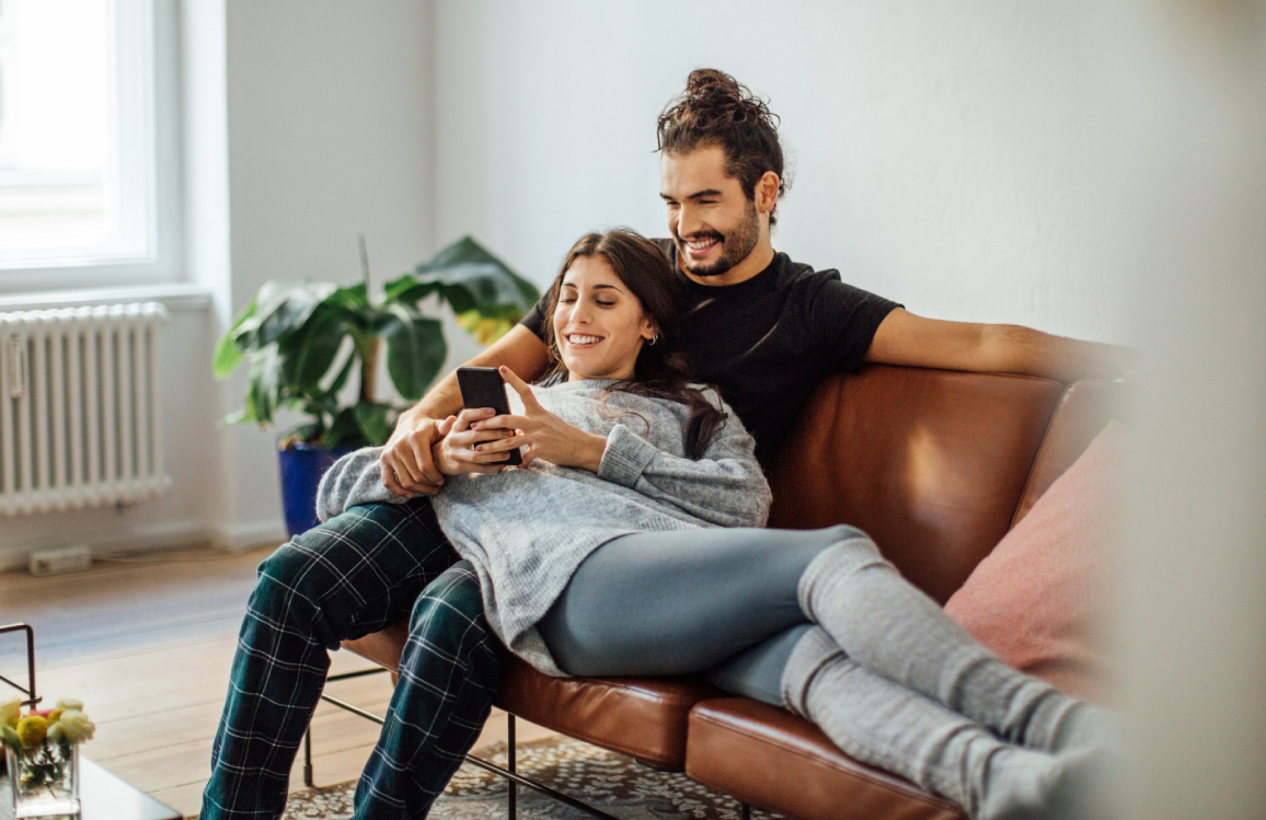 Ein Pärchen sitzt bzw. liegt auf dem Sofa und beide schauen gemeinsam auf ein Smartphone.