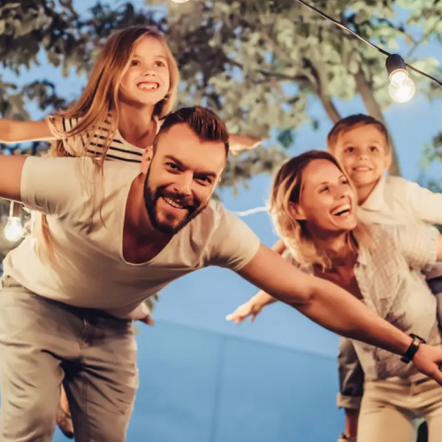 Eine Familie steht im Freien. Im Hintergrund ist eine Leuchtgirlande zu sehen. Die Kinder tun so, als würden sie auf dem Rücken der Eltern fliegen.
