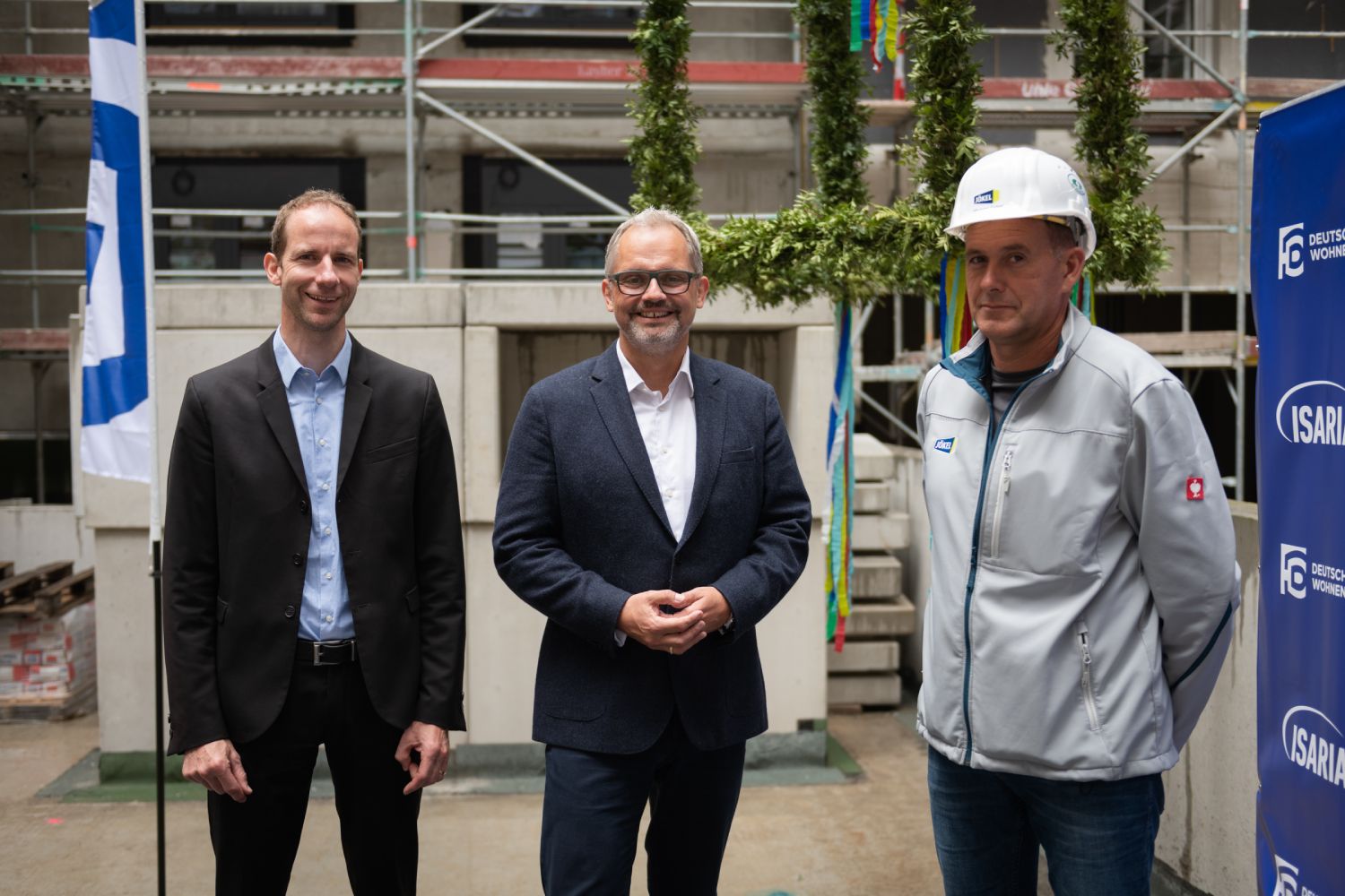 Drei Männer stehen auf einer Baustelle vor einem Richtkranz. Zwei sind in Anzügen gekleidet, der Andere casual mit einem Bauhelm. Sie lächeln in die Kamera.