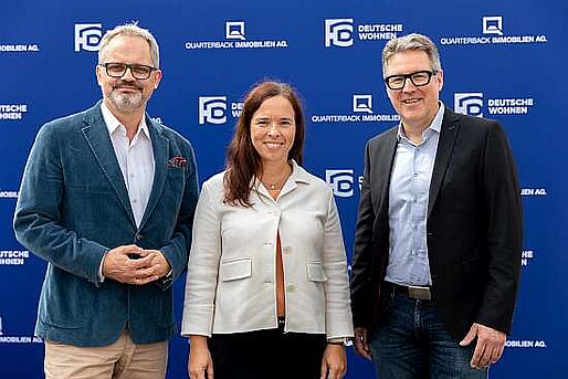 Eine Frau geflankt von zwei Männern stehen vor einer blauen Wand mit dem Logo der Deutsche Wohnen. Alle sind schick angezogen und lächeln in die Kamera.