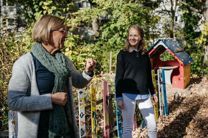 Zwei Frauen unterhalten sich drau&szlig;en in einem Garten neben einem bunt bemaltem Zaun und einem Spielh&auml;uschen f&uuml;r Kinder.