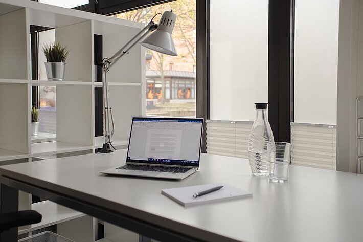 Ein Tisch im Coworking Space auf dem ein Laptop, eine Flasche Wasser und ein Glas sowie ein Notizblock mit Stift liegt. Neben dem Tisch steht ein Regal.