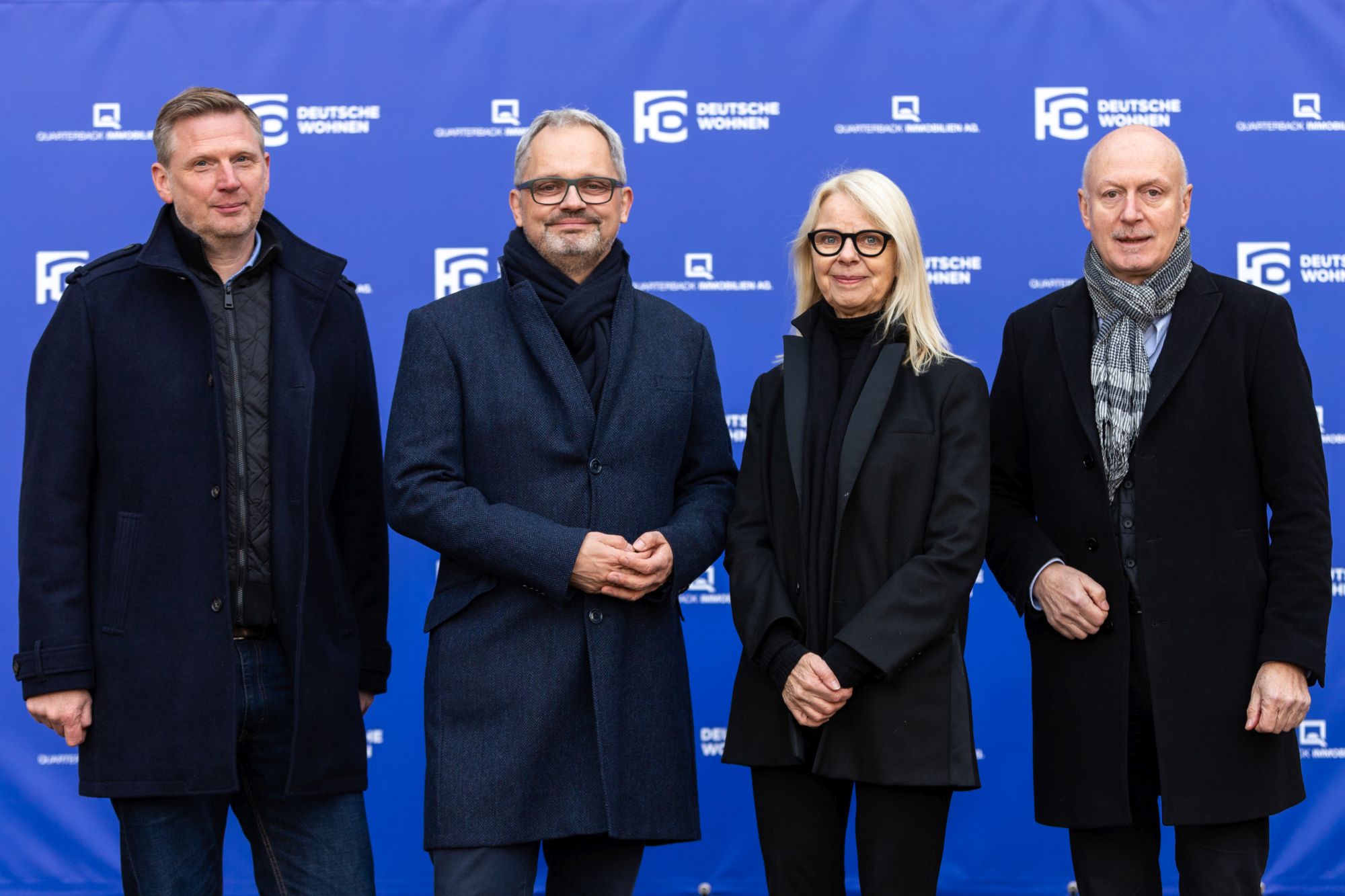 Drei Männer und eine Frau stehen vor einem blauen Hintergrund mit dem Deutsche Wohnen Logo. Sie sind bedeckt und schick gekleidet und lächeln in die Kamera.