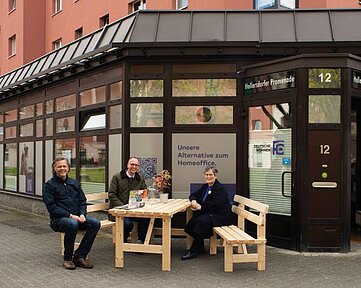 Drei Menschen sitzen auf auf Bänken an einem Tisch vor einem Gebäude. Auf dem Gebäude ist das Logo der Deutsche Wohnen sowie der Satz „Unsere Alternative zum Home Office“.