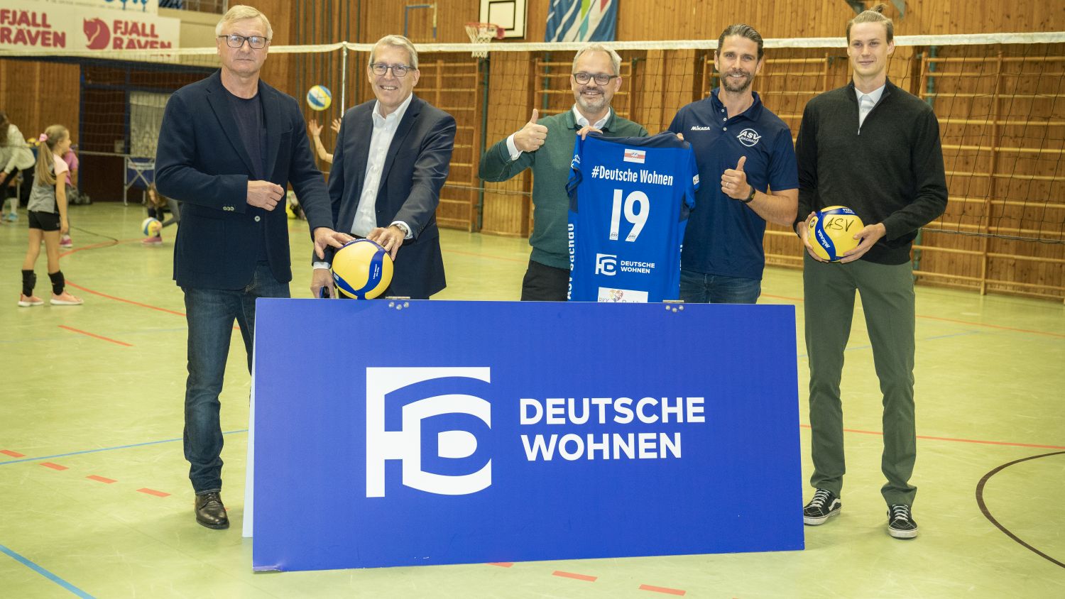5 Männer stehen in einer Sporthalle vor einem Deutsche Wohnen Aufsteller. Sie halten Volleybälle und ein Trikot, auf welchem die Deutsche Wohnen als Sponsor drauf ist.